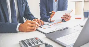 Dupla de profissionais em traje de negócios discutindo gráficos e relatórios em uma mesa, com computador e calculadora ao fundo.